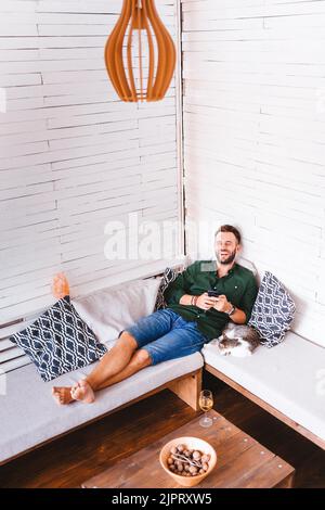 A happy young male with his cat using his mobile phone on the sofa at home Stock Photo