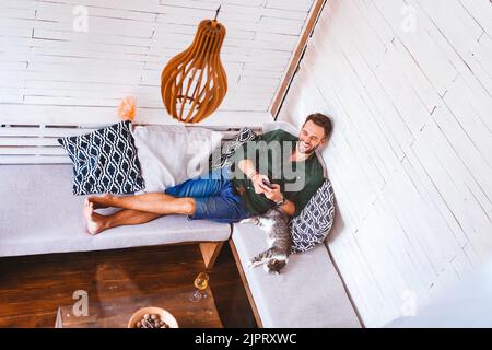A happy young male with his cat using his mobile phone on the sofa at home Stock Photo