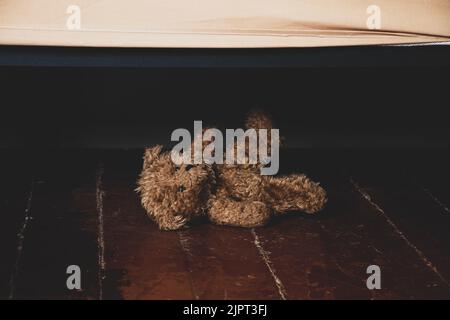 by the bed on the floor in the bedroom of the house lies a teddy brown bear, a teddy bear under the bed on the floor in the dark Stock Photo
