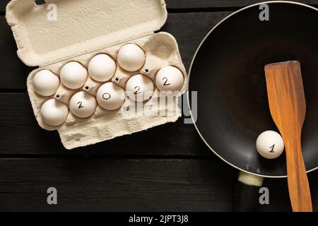 chicken eggs in boxes and text 2022 on eggs, happy new year 2022, chicken eggs on the table and frying pan, fry scrambled eggs Stock Photo