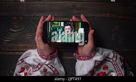 August 2022 - Kyiv, Ukraine. Watching online speech of President Volodymyr Zelenskiy on smartphone on wooden table background. News from front Stock Photo