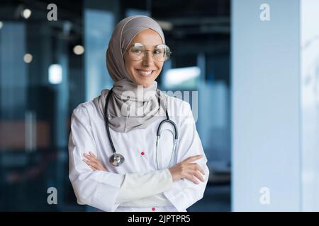 Portrait of Arabic female doctor in modern clinic, Muslim woman wearing hijab glasses and white medical coat working in hospital office, smiling and looking at camera with crossed arms Stock Photo