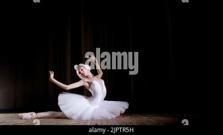 in rays of spotlight, on the stage of the old theater hall. Young ballerina in suit of white swan and pointe shoes, dances elegantly certain ballet motion, Swan Lake. High quality photo Stock Photo
