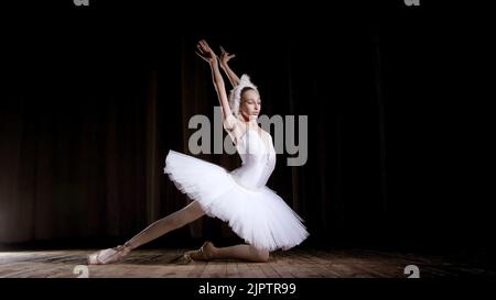 in rays of spotlight, on the stage of the old theater hall. Young ballerina in suit of white swan and pointe shoes, dances elegantly certain ballet motion, part de bras. High quality photo Stock Photo