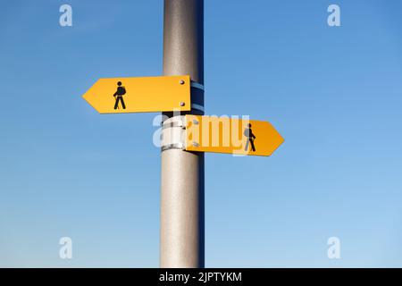 two yellow hiking trail signs with symbol person, backpack, hiking sticks, the signs point in different directions, in front of blue cloudless sky Stock Photo