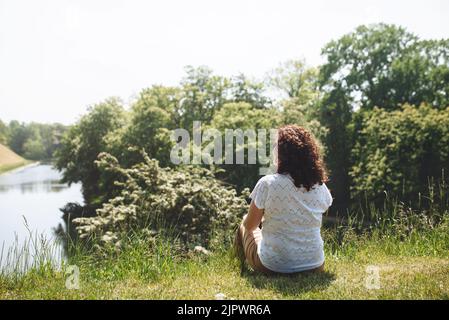 Back view Girl traveler sitting on green grass and looking at lake and trees. Woman with curly hair. The simple pleasure of solitary silence. Digital detox. Copy Space for text. Stock Photo