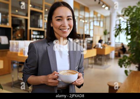Smiling young Asian professional woman wearing suit holding coffee, portrait Stock Photo