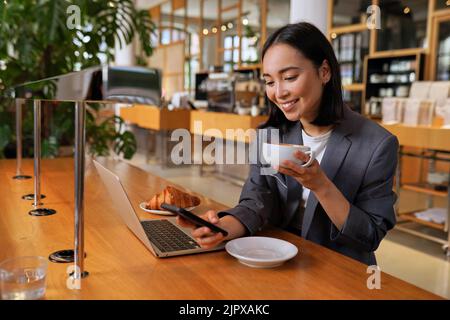 Young Asian business woman wearing suit drinking coffee using phone in cafe. Stock Photo