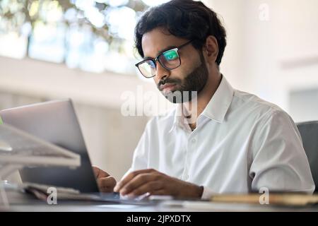 Busy indian business man employee working typing on laptop sitting at desk. Stock Photo