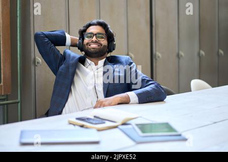 Young happy indian business man wearing headphones sitting at workplace. Stock Photo