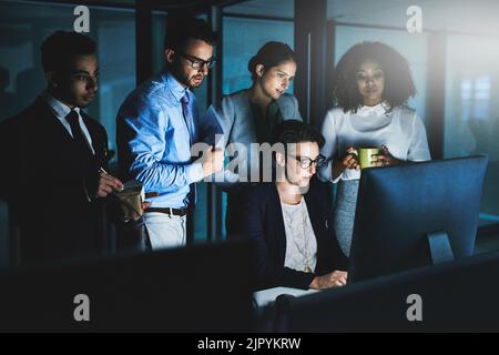 Getting the job done in limited time with unlimited resources. colleagues standing together as they work on something on a computer at night. Stock Photo
