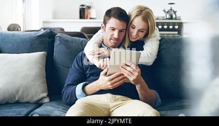 What you working on. an affectionate young couple using a digital tablet while sitting on their sofa at home. Stock Photo