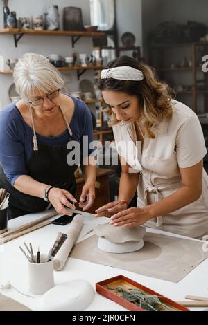 People working with clay on the table. Woman on pottery workshop in studio. Adults learning to do ceramic plates. Pottery as hobby and leisure Stock Photo