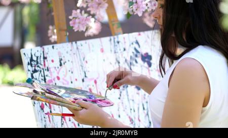 a beautiful woman painter in white dress, in blooming spring apple orchard, she mixes paints with special small spatula on the pallet. artist paints a picture of flowers. High quality photo Stock Photo
