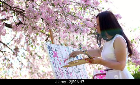 a beautiful woman painter in white dress, artist paints a picture of flowers in blooming spring apple orchard, she applies paints to the canvas with a special small spatula, using a special drawing technique. holds a palette with paints. High quality photo Stock Photo