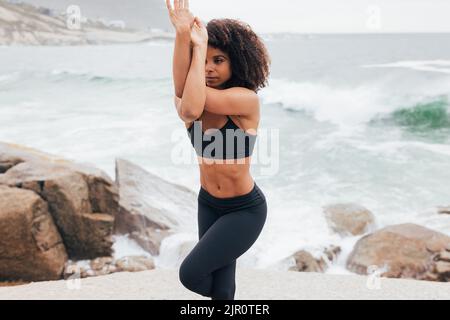 Woman practicing Eagle pose near an ocean. Young female doing yoga training at sea shore. Stock Photo