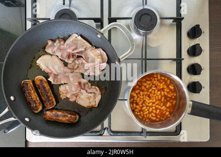 Traditional English breakfast ingredients cooking and frying on a gas hob. Bacon, sausages and baked beans. UK. Concept - unhealthy eating Stock Photo