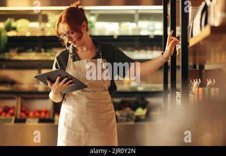 Female supermarket owner using a digital tablet while standing in her shop. Young female entrepreneur running her small business using wireless techno Stock Photo