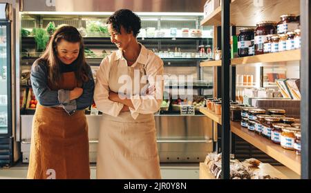 Two grocery store workers laughing happily while standing together in their shop. Cheerful woman with Down syndrome working in a local supermarket wit Stock Photo