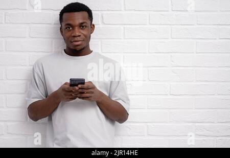 Black young man using smartphone over white brick background, finger, reading social media internet, typing text or shopping online, copyspace . Stock Photo