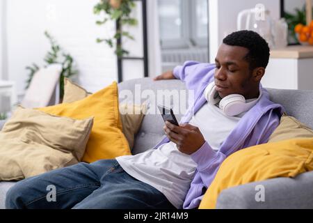 Black young man using smartphone at home sitting on couch, pressing finger, reading social media internet, typing text or shopping online, Making Stock Photo