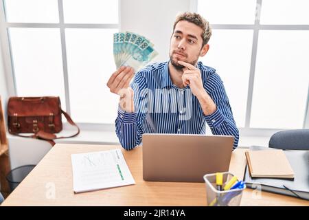 Hispanic man with beard working at the office holding brazilian reals serious face thinking about question with hand on chin, thoughtful about confusi Stock Photo