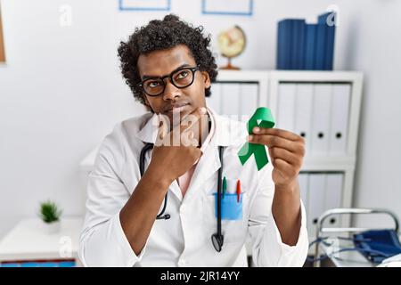 African doctor man holding support green ribbon serious face thinking about question with hand on chin, thoughtful about confusing idea Stock Photo