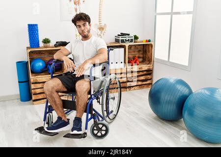 Hispanic man sitting on wheelchair at physiotherapy clinic making fish face with lips, crazy and comical gesture. funny expression. Stock Photo