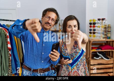 Middle age interracial couple at retail shop using smartphone looking unhappy and angry showing rejection and negative with thumbs down gesture. bad e Stock Photo