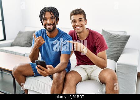 Young hispanic men using smartphone sitting on the sofa at home pointing to the back behind with hand and thumbs up, smiling confident Stock Photo