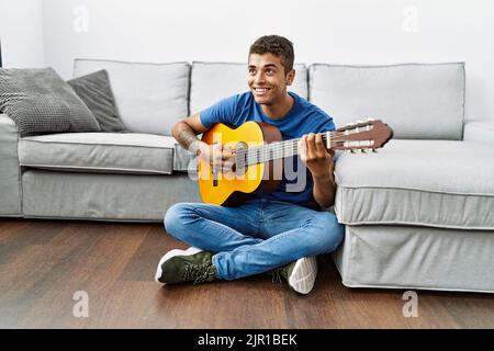 Young hispanic man playing guitar sitting on the floor at home Stock Photo