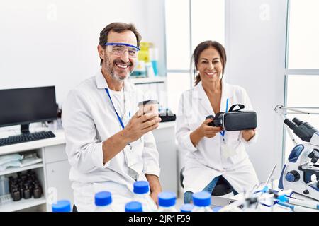 Middle age man and woman partners wearing scientist uniform drinking coffee and holding vr goggles at laboratory Stock Photo