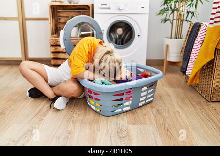 Young blonde girl doing laundry putting clothes into washing machine at home. Stock Photo