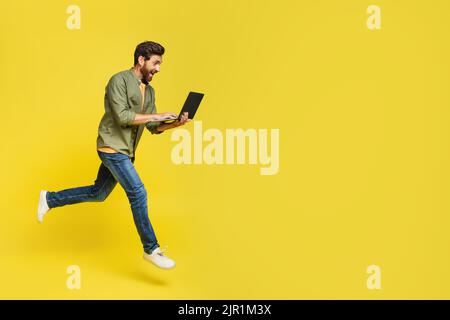 Full length of excited middle aged man using laptop computer and jumping on yellow background copy space Stock Photo