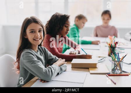 Happy Schoolgirl Sitting At Desk Smiling To Camera In Classroom Stock Photo