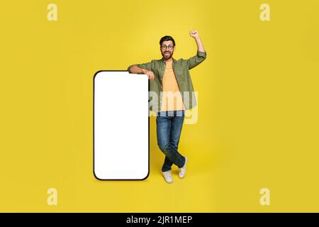 Excited man leaning on huge cellphone with blank screen and shaking fist in joy, yellow studio background, mockup Stock Photo