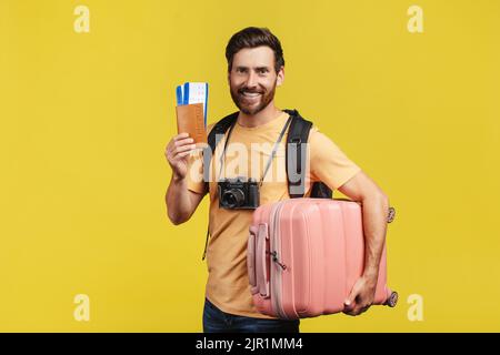 Vacation concept. Excited male tourist wearing backpack, holding suitcase, passport with tickets over yellow background Stock Photo
