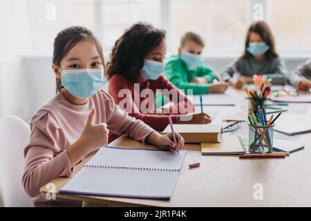 Asian Girl Gesturing Thumbs Up Wearing Face Mask In Classroom Stock Photo