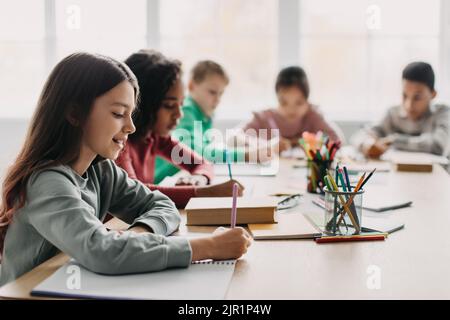 Schoolgirl Writing Essay During Class With Mixed Children In Classroom Stock Photo