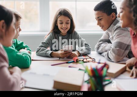 Cheerful Diverse School Children Using Cellphone Sitting In Modern Classroom Stock Photo