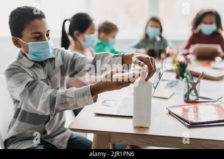 Black Schoolboy Disinfecting Hands With Sanitizer Sitting In Modern Classroom Stock Photo