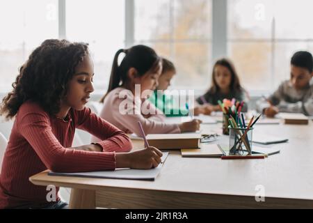 African Schoolgirl Taking Notes Sitting With Diverse Classmates In Classroom Stock Photo