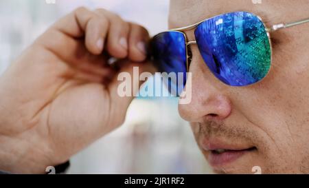 close-up, portrait of Handsome young man trying on blue sunglasses at optical store, optician retail store. in glasses of spectacles is reflected the showcase of optics with glasses. High quality photo Stock Photo