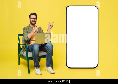Excited middle aged man sitting on chair using laptop pc and pointing at big cellphone with empty white screen Stock Photo