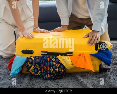Yellow suitcase with couple lovers hands trying to close it with full colorful of many clothes, packing bag together. Preparing to vacation. Couple tr