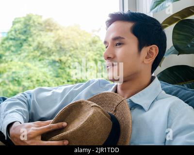 Portrait of handsome Asian man in light blue denim shirt and white tee holding brown beach hat sitting alone on cozy sofa seat near glass window and g Stock Photo