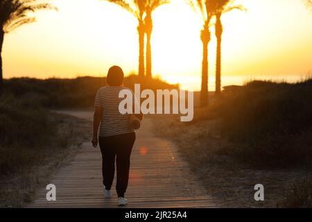 back view of active overweight woman with yoga mat walking to beach to practice yoga, exercising outdoors on sunrise in the morning. Concept of health Stock Photo