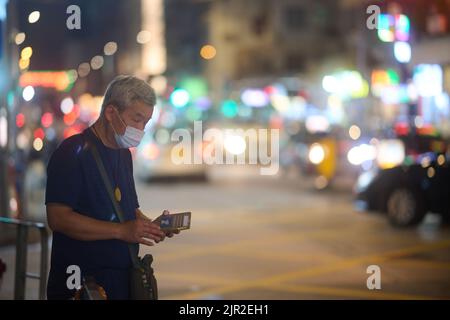 A senior asian man in a mask using mobile phone near a road in Kowloon City, Hong Kong Stock Photo