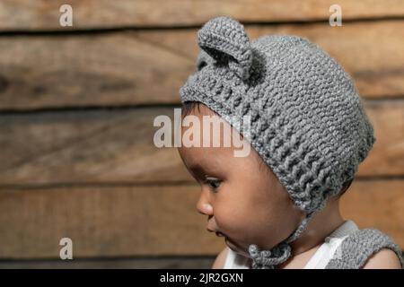 beautiful baby latina with brown skin, distracted looking to one side, with her face in profile showing her left side, crochet cap on a wooden backgro Stock Photo