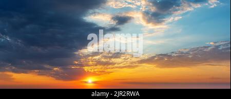 Setting sun in the orange blue evening sky. Sun rays break through the light clouds at sunset. Dark stormy cloud covering half the sky at sundown. Stock Photo